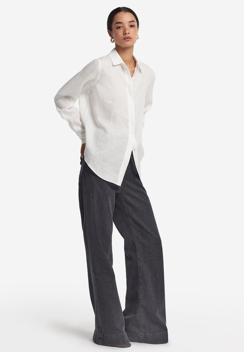 Camicia bianca in lino con bottoni e colletto, indossata con pantaloni larghi in denim scuro. La camicia presenta una vestibilità rilassata e un orlo curvo.
