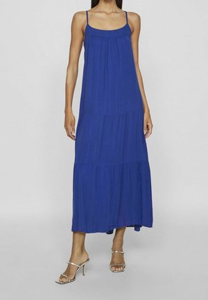 Kobieta ubrana w bezrękawnikową, warstwową, królewską niebieską suknię maxi, zestawioną ze srebrnymi sandałami na wysokim obcasie, na jednolitym tle.