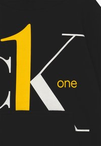 Felpa nera con grande testo grafico bianco e giallo "CK1 one" dal design moderno. Tessuto morbido, dettaglio con tasca frontale.