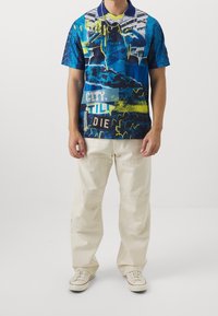 Polo tričko s krátkým rukávem v modrožlutém provedení s grafickou koláží. Má jednobarevný modrý límec a krátké rukávy, v kombinaci s světle béžovými kalhotami.