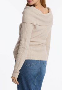 Beige Ripp-Pullover mit weitem, schulterfreien Ausschnitt und enganliegenden Ärmeln, kombiniert mit blauer Denim. Dezente Textur im gesamten Design.
