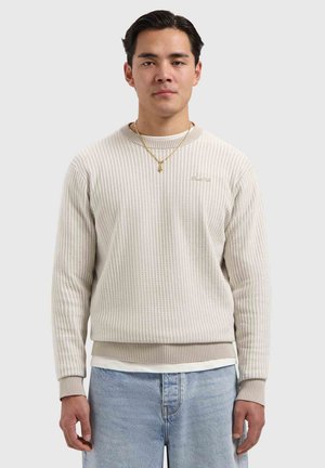 Jonge man die een beige geribbelde trui draagt over een wit overhemd, lichtblauwe spijkerbroek en een gouden ketting, staand tegen een effen achtergrond.