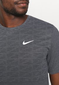 Szary koszulka sportowa z teksturowanym wzorem, krótki rękaw i białe logo Nike na lewej części klatki piersiowej.