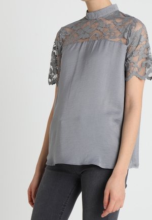 Femme portant un chemisier gris avec des manches courtes en dentelle et un empiècement, associé à un pantalon gris foncé ajusté.