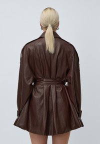 Blondīne ar astīti,ģērbta tumši brūnā ādas jakā ar jostu, skatīta no aizmugures uz vienkārša gaiša fona.
