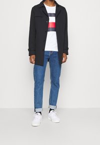 Abrigo negro con cuello, cierre de botones; camisa blanca con rayas horizontales rojas y azul marino; jeans azules de pierna recta; zapatillas blancas.