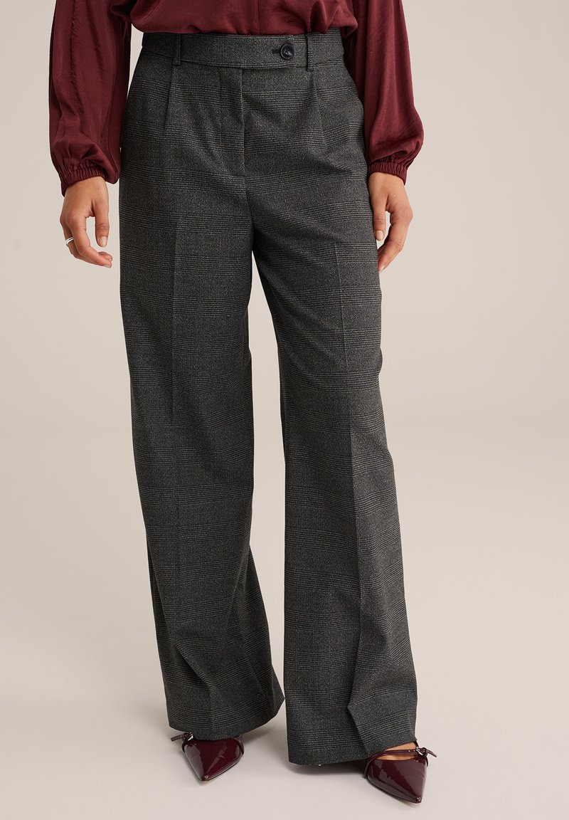 Donna che indossa pantaloni larghi a quadri grigi, una camicia bordeaux a maniche lunghe, e scarpe con punta bordeaux in un ambiente neutro.
