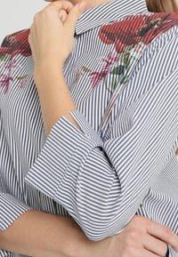 Kobieta w paskowanej koszuli na guziki z kwiatową haftem, trzymająca kołnierzyk jedną ręką i skrzyżowane ręce.