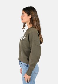 Sweat-shirt vert olive avec un col rond, des épaules tombantes et du texte brodé blanc. Associé à un jean en denim bleu clair.
