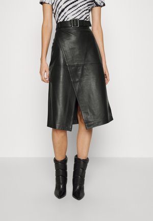Femme portant une jupe midi en similicuir noire ceinturée, avec une fente asymétrique sur le devant et des bottines noires matelassées.