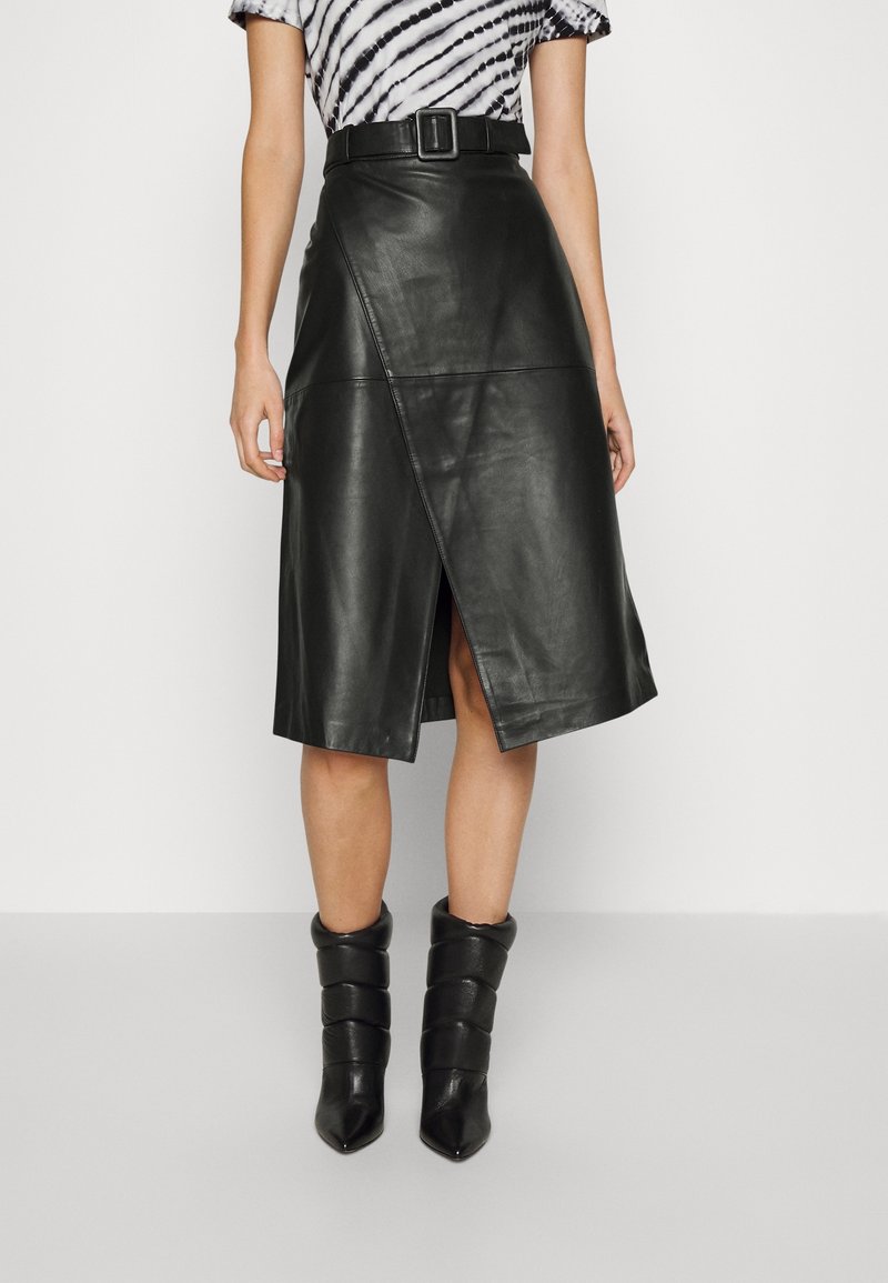 Femme portant une jupe midi en similicuir noire ceinturée, avec une fente asymétrique sur le devant et des bottines noires matelassées.