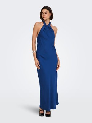 Femeie purtând o rochie halter fără mâneci, până în pământ, de culoare albastru royal, cu pantofi cu toc negri și o brățară argintie, stând în fața unui fundal alb.