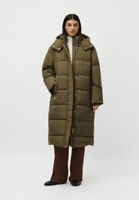 Woman wearing a long olive green padded coat with hood, cream sweater, brown pants, and black shoes standing against a white background.