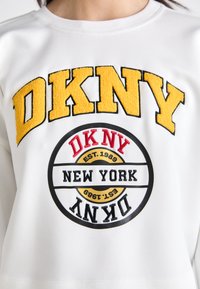 Λευκό φούτερ με το "DKNY" σε ανάγλυφα κίτρινα γράμματα και ένα κυκλικό λογότυπο σε μαύρο και κόκκινο χρώμα που περιλαμβάνει το "NEW YORK" και ημερομηνίες ίδρυσης.