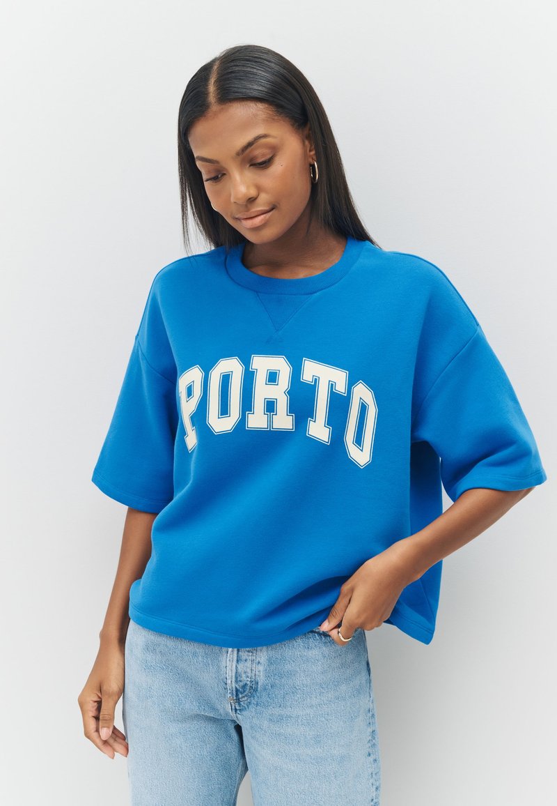 Ķēnišķīgi zils pārlielgabarīta sporta krekls ar uzdrukātu vārdu "PORTO" baltā krāsā, apaļu kakla izgriezumu un īsām piedurknēm, kopā ar gaiši zilām džinsa biksēm.
