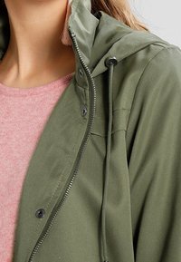 Veste à capuche vert olive avec fermeture éclair et boutons à pression, associée à une chemise rose pâle. La veste présente une texture lisse et une capuche ajustable.