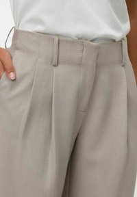 Svetlo bež hlače po meri iz gladkega blaga, z naborki, ravnim sprednjim delom in stranskimi žepi za strukturiran videz.