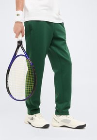 Άτομο με πράσινο παντελόνι, λευκά παπούτσια Lacoste και βραχιόλι κρατά μπλε και κίτρινη ρακέτα τένις στο πλάι.