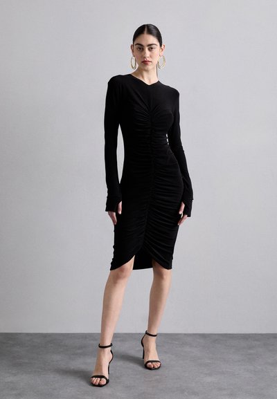 Black long-sleeve dress with ruched detailing and a curved hem. Made of soft fabric; paired with black strappy heels. Solid color throughout.