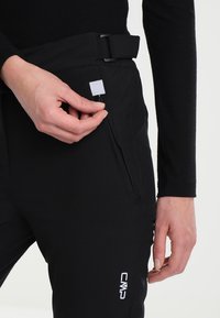 Black softshell pants with a zippered pocket, white label, and embroidered logo. Features a belted waistband and smooth texture.