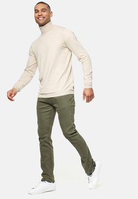 Jersey de cuello alto beige en material suave, combinado con pantalones ajustados verdes oliva. La modelo lleva zapatillas blancas, caminando con confianza.