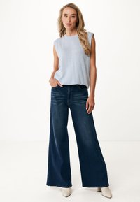 Jasnoniebieski top bez rękawów z okrągłym dekoltem, w zestawieniu z szerokimi ciemnoniebieskimi dżinsami. Modelka stoi w beżowych szpilkach, z rękami w kieszeniach.