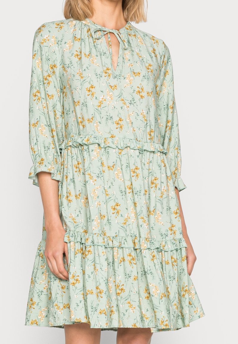 Robe florale vert clair avec des fleurs jaunes et blanches, manches trois-quarts, volants superposés et décolleté goutte d'eau avec un nœud.