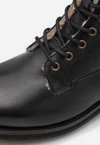 Bota de cuero negra con un acabado suave, que presenta ojales de latón, cordones negros y un forro de tela texturizada. Suela robusta con detalle de costura.