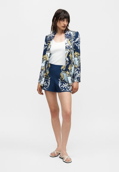 Γυναίκα που φοράει ναυτικό μπλέ σακάκι και αντίστοιχα σορτς με λευκά και χρυσά διακοσμητικά σχέδια, λευκό αμάνικο τοπ και λευκά σανδάλια με λουριά.