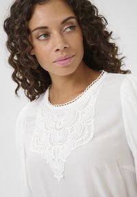 Blusa bianca con dettagli in pizzo intricati sul collo, tessuto morbido e maniche a sbuffo per un aspetto delicato.