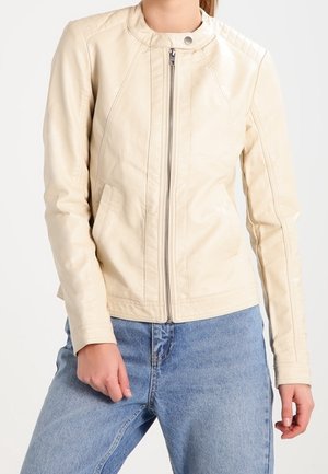 Beige zip-up leather jacket with stand collar and snap button, worn with light blue jeans, hands at sides and front pocket detail visible.