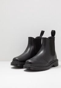 Botas Chelsea negras con paneles laterales elásticos y tiradores, reposando sobre una superficie blanca con un fondo gris claro.