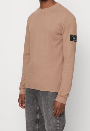 Man draagt een beige geribde trui met lange mouwen met een Calvin Klein logo patch op de linkerarm, gecombineerd met grijs gewassen spijkerbroek.