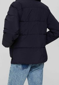 Person iført en sort pufferjakke og lyseblå jeans, vist fra ryggen med hænderne stukket ind i jakke-lommerne.