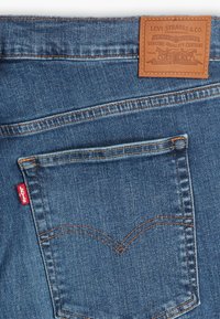 Denim-farkut, joissa on vaaleansininen pesu, kontrastiväriset ompeleet, nahkainen takapläntti brändilogoineen ja pieni punainen brändilappu taskussa.