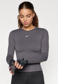 Športna majica z dolgimi rokavi temno siva z okroglim izrezom in belim logotipom Nike. Model nosi črne brezprstne rokavice s teksturiranim prijemom.