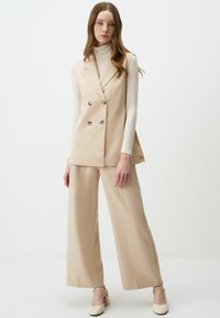 Beige dubbele-breasted vest met grote knopen, gedragen over een lichte turtleneck en brede beige broek; gladde stof en slim fit.