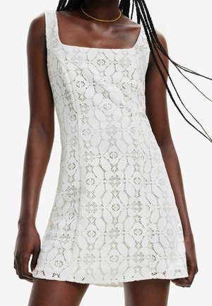 Kobieta w białej sukience bez rękawów z koronki, o kwadratowym dekolcie, w kwiatowy wzór, z dołem sięgającym powyżej kolan.