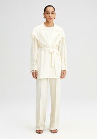 Prenda exterior de color crema, texturada, con cuello chal y detalles de flecos, combinada con pantalones de pierna ancha a juego y sandalias con tiras.