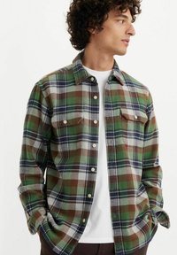 Camisa de franela con un patrón de cuadros en verde, marrón y gris, mangas largas, dos bolsillos en el pecho, diseño de botones en la parte delantera y una textura suave.
