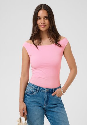 Woman with long brown hair wearing pink off-shoulder top and blue jeans, holding sunglasses in left hand, right hand in pocket.