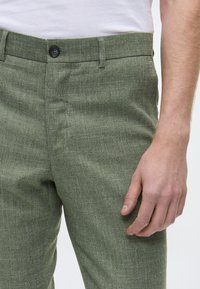 Grüne strukturierte Hose aus gewebtem Stoff, mit einem Knopfverschluss und Gürtelschlaufen, präsentiert mit einer Hand, die in der Nähe der Taille ruht.