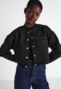 Čierna skrátená bomber bunda vyrobená z hladkého materiálu. Obsahuje strieborné cvoky, malú náprsné vrecko a okrúhly golier.