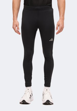 Mężczyzna ubrany w czarne przylegające legginsy sportowe i białe buty sportowe Adidas, stojący z ramionami zrelaksowanymi wzdłuż ciała.