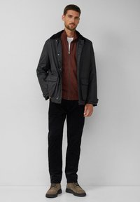 Schwarze Herrenjacke mit Kragen, darüber getragen ein burgunderrotes, geripptes Zip-up-Oberteil, kombiniert mit schwarzen Hosen und beigen Stiefeln. Glatte Textur, lässige Passform.