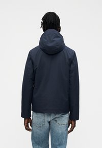 Mand med dreadlocks iført en marineblå hættetrøje og lyseblå jeans, set bagfra mod en ensartet hvid baggrund.