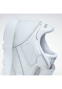 Zapatilla deportiva blanca con una parte superior sintética texturizada, detalles cosidos, cuello acolchado y suela de goma. Presenta un logo en el lateral.