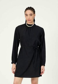 Γυναίκα με πλεξούδα, ντυμένη με μαύρο φόρεμα μακρυμάνικο με ζωνάκι στη μέση και πολύχρωμο κολιέ από χάντρες, που στέκεται μπροστά σε απλό φόντο.