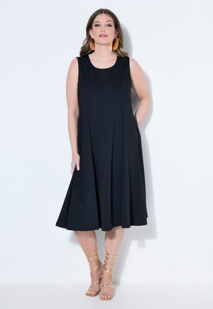 Kvinde iført sort ærmeløs midikjole med flæset nederdel, brune dråbeøreringe og gyldne remsandaletter med høje hæle, stående op ad en hvid væg.