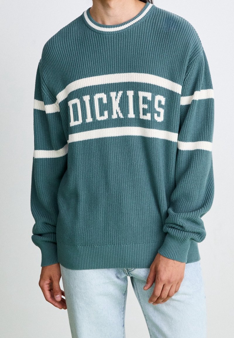 Personne portant un pull en maille bleu sarcelle avec des rayures blanches et le texte « DICKIES » sur la poitrine, associé à un jean bleu clair.